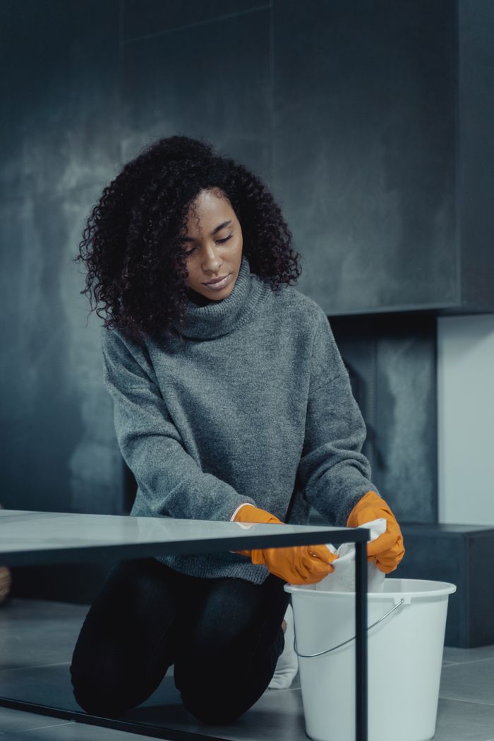 hero-img-01 African American woman in gray sweater cleaning with a pail and gloves at home.